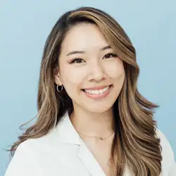 Professional headshot of a person with long wavy hair, wearing a white coat and small hoop earrings, posed against a light blue background.